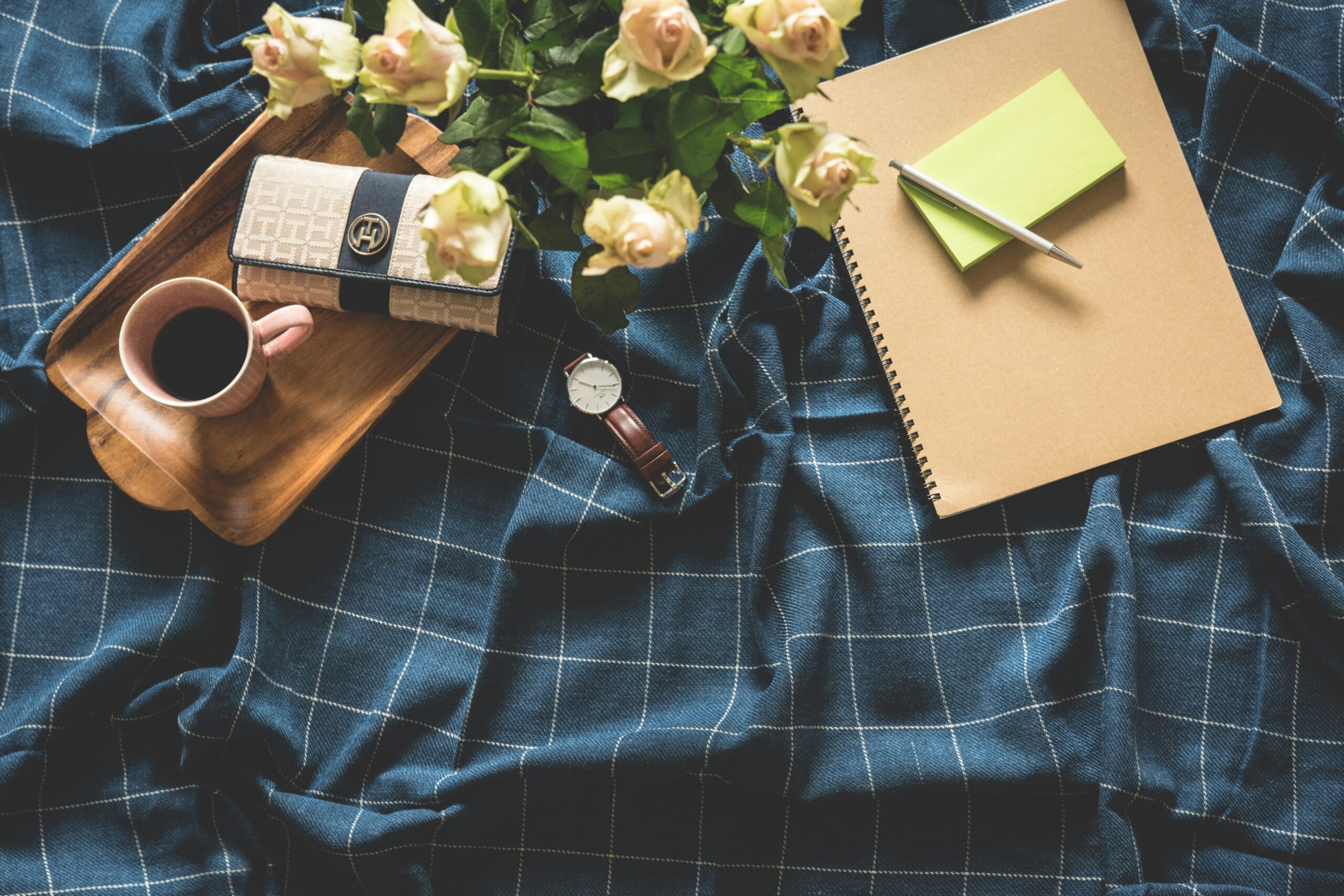 A cozy flatlay showing coffee, roses, notebook, and watch on a checkered blanket.